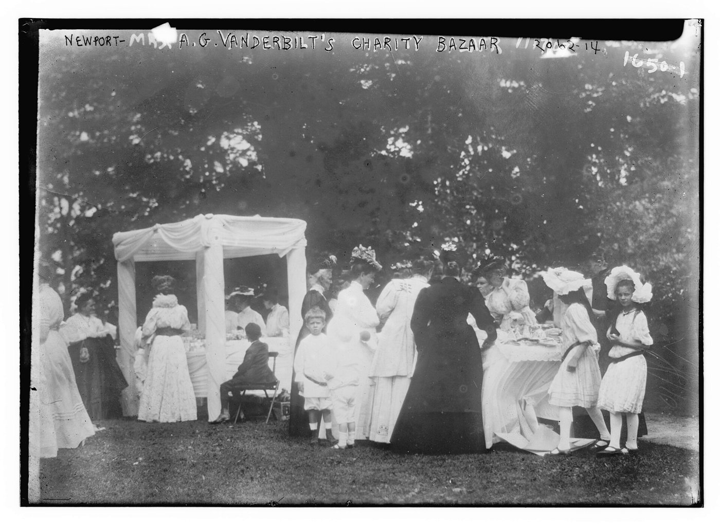 Fotografía antigua en blanco y negro de un bazar benéfico en un jardín, con mujeres y niñas vestidas de blanco ante un puesto
